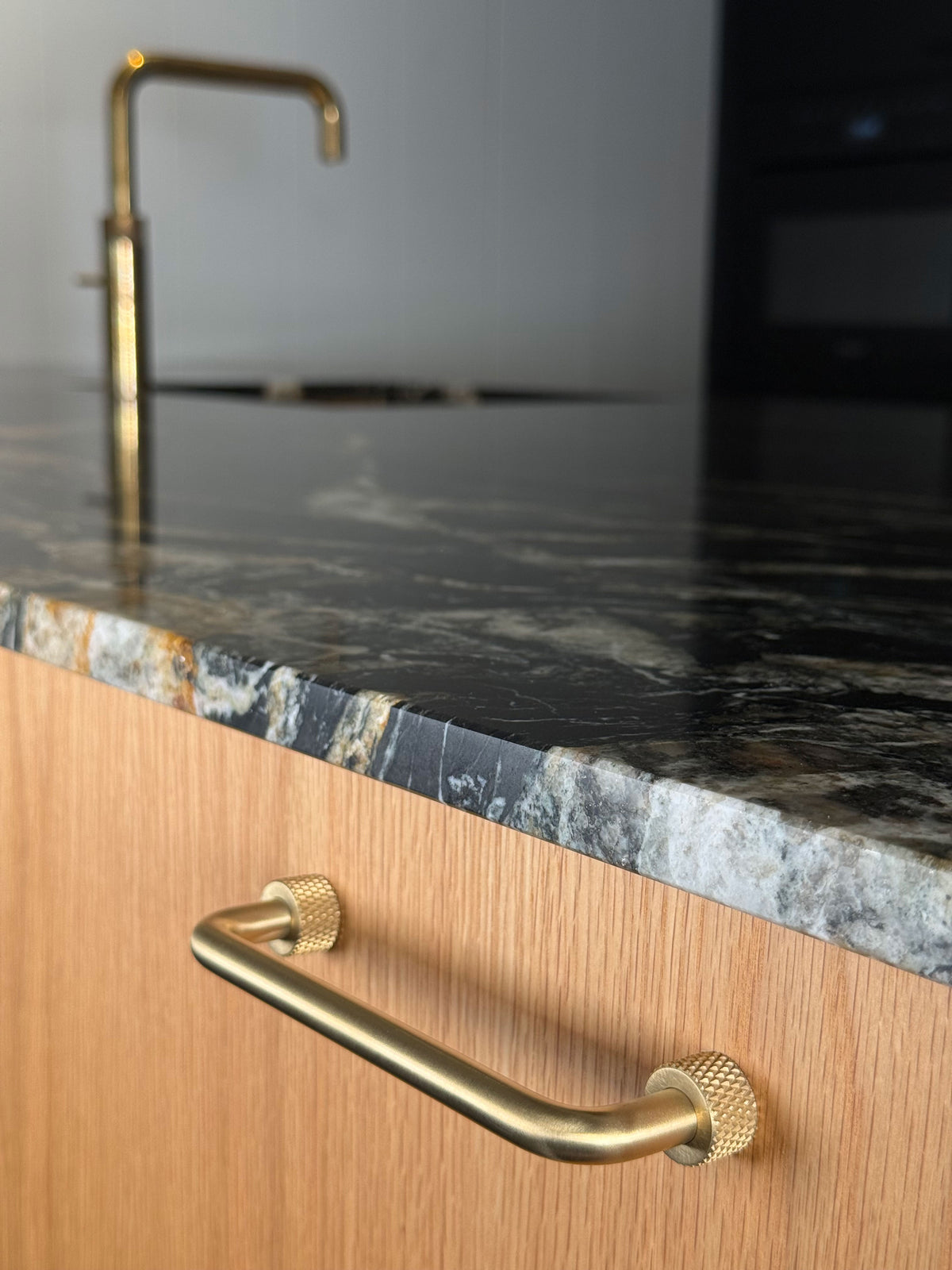 Trianglen handle in unlacquered brushed brass on a wooden cabinet front below a dark worktop with a kitchen tap