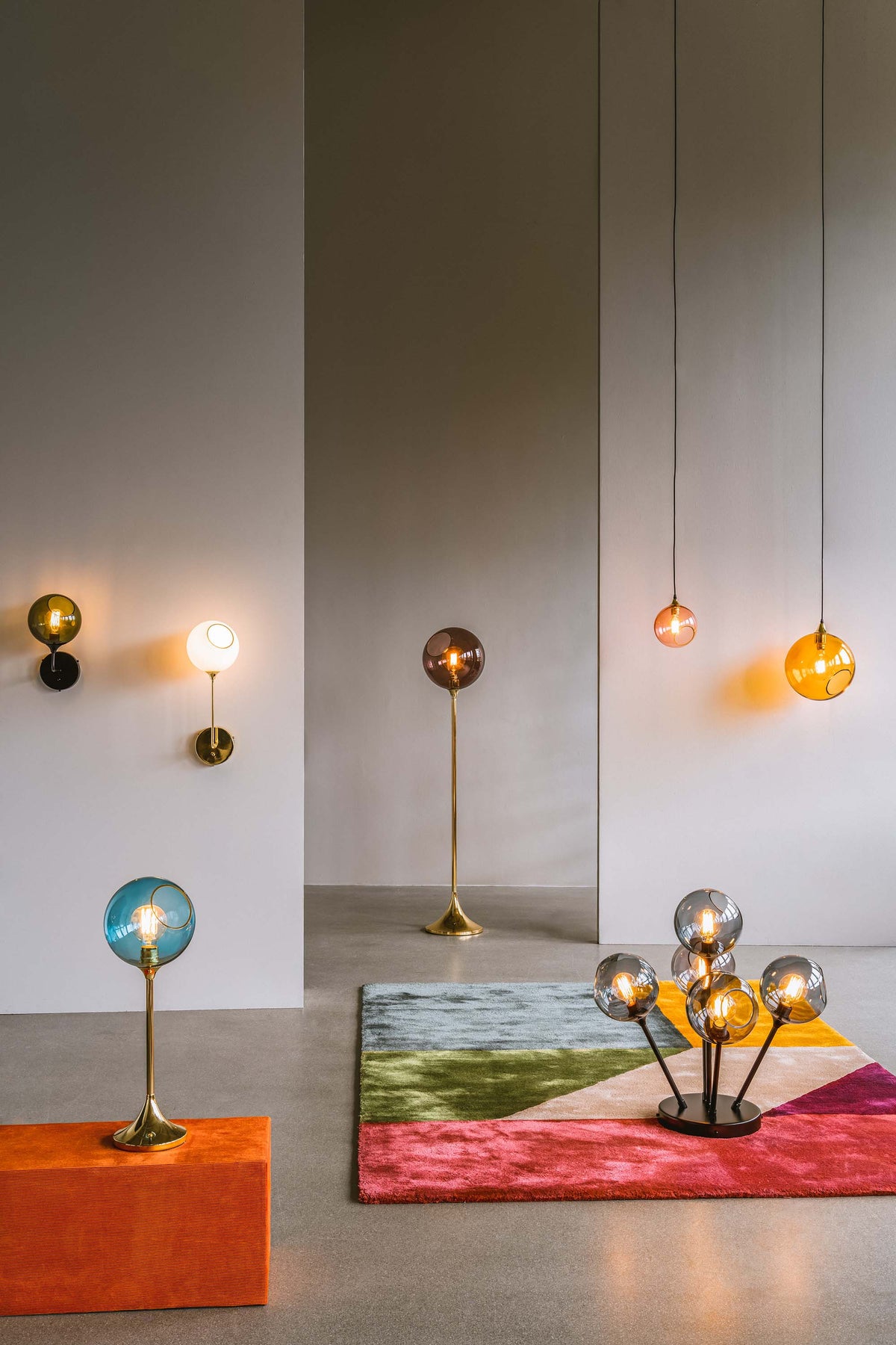 Rund lampe i orange/gult glas og med et afskåret stykke med guldkant og kig til pæren. Fatningen og rosetten er guldfarvet og ledningen er af sort stof.