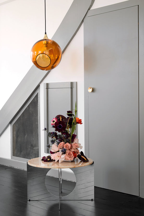 Rund lampe i orange/gult glas og med et afskåret stykke med guldkant og kig til pæren. Fatningen og rosetten er guldfarvet og ledningen er af sort stof.