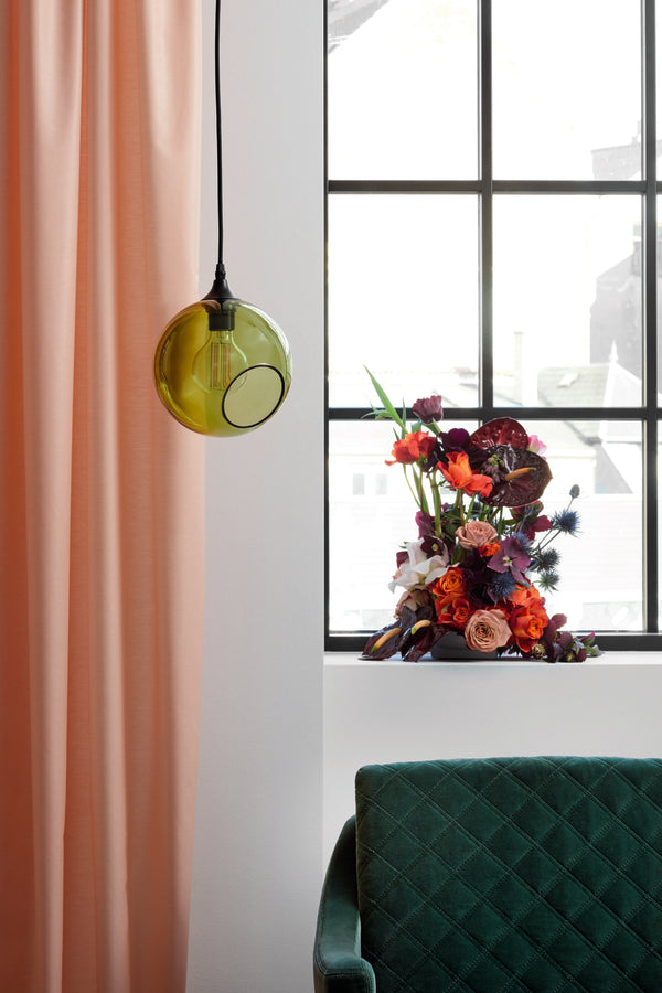 Rund lampe i grøn glas med et afskåret stykke med kig til pæren. Fatning er sort og ledningen er af sort stof.